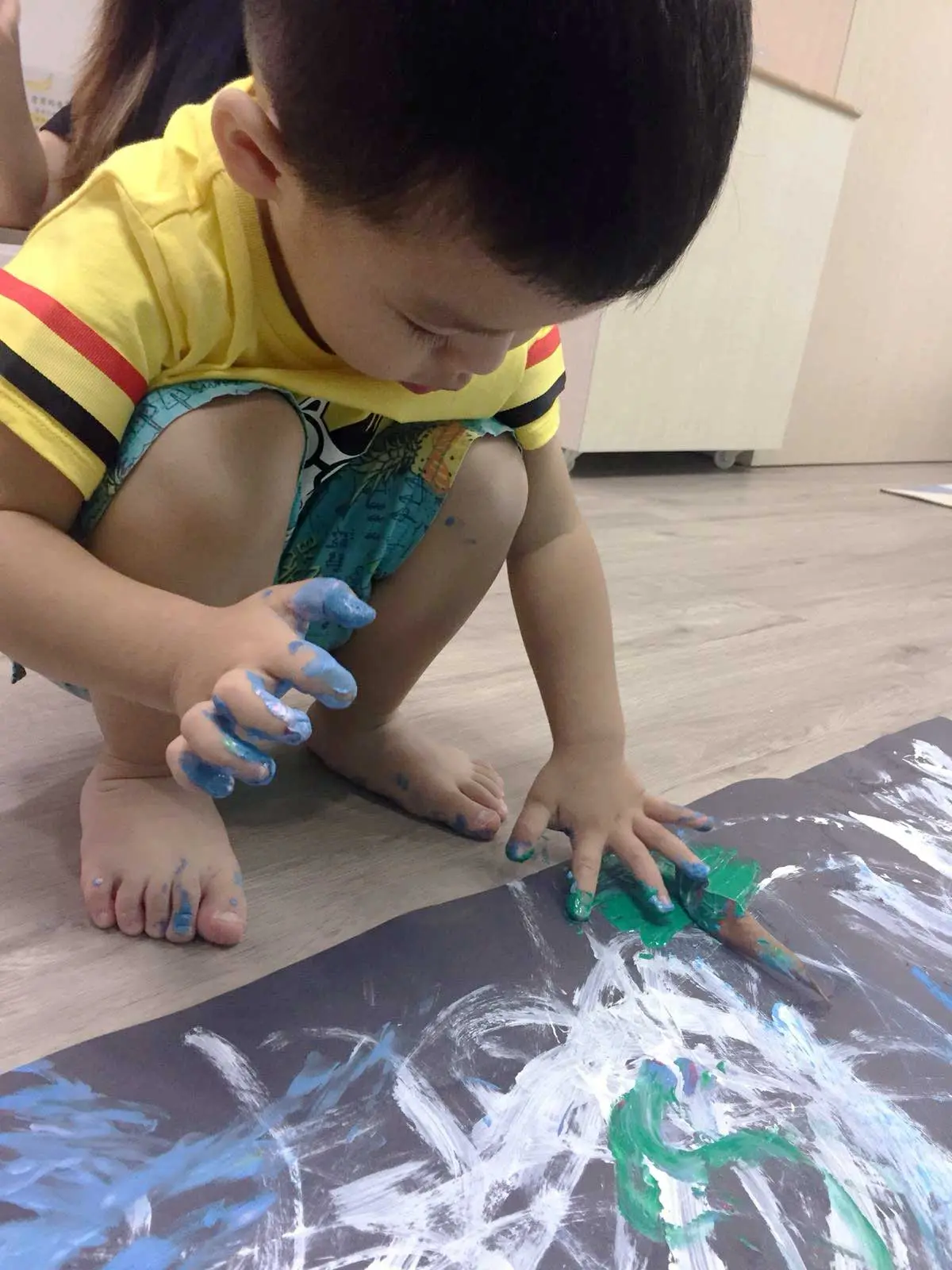 青と緑の絵の具を指で広げる幼い子ども。