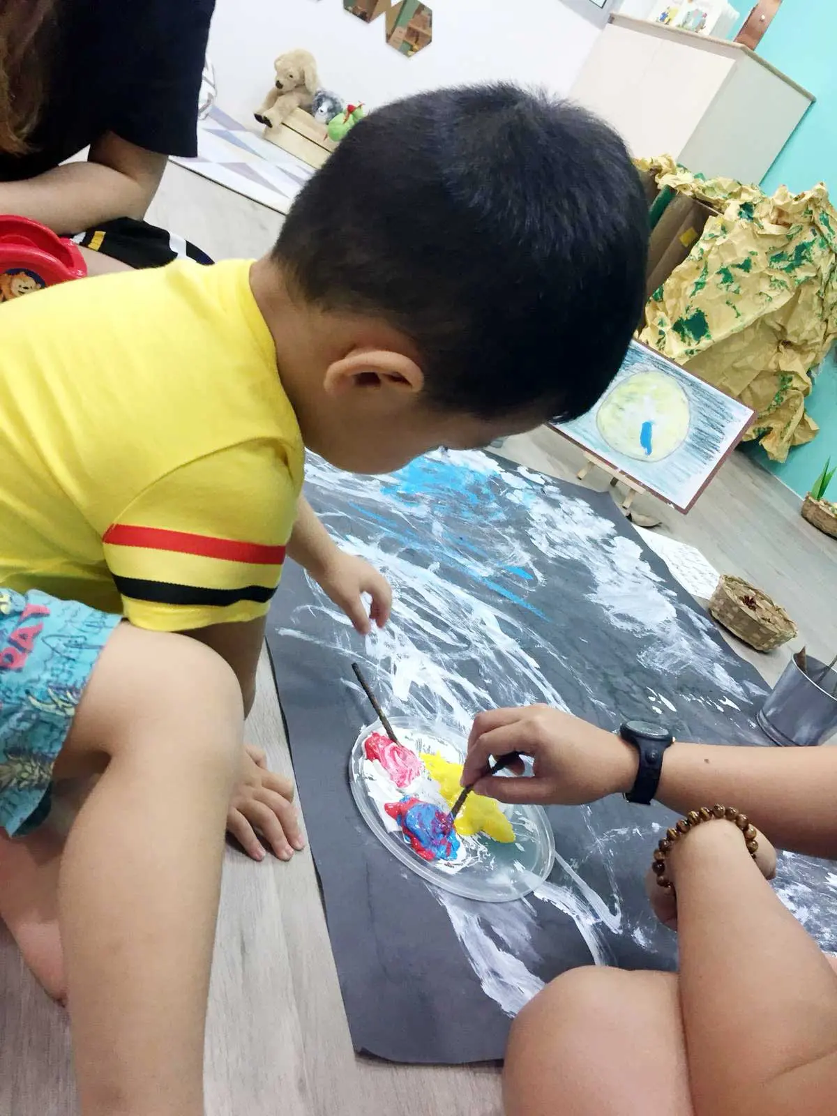 大きな作品のそばで、大人が絵の具を混ぜるのを見る子ども。